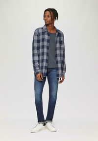 Chemise écossaise navy et grise, t-shirt gris, jeans en denim et baskets blanches ; coupe décontractée et texture lisse avec fermeture à boutons et poche unique.