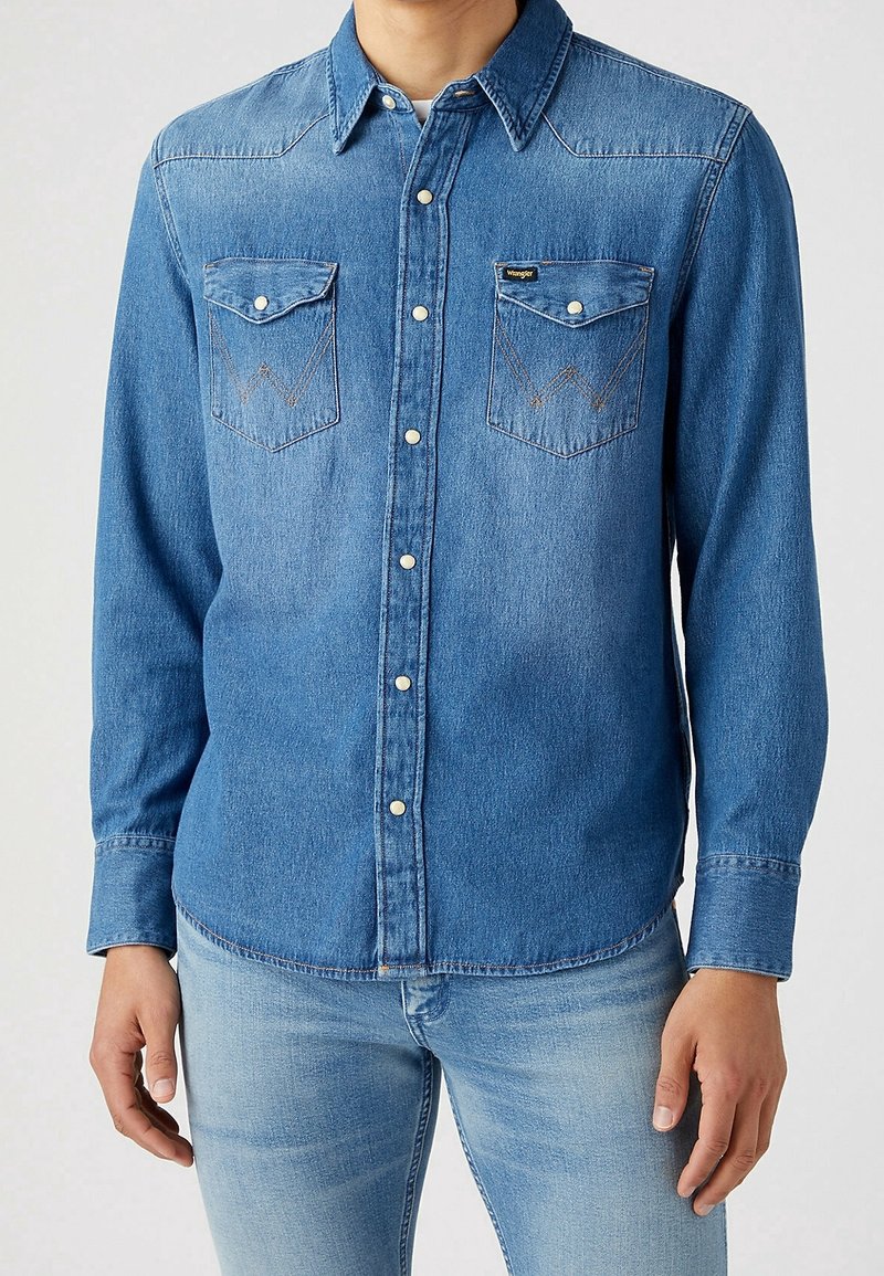 Jasnoniebieska koszula dżinsowa z kołnierzykiem zapinanym na guziki, dwoma kieszeniami na piersi oraz kontrastującymi szwami w wzór zygzakowy. Gładka faktura.