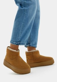 Brown suede ankle boots with a soft fleece lining, rounded design, and thick rubber soles, paired with cuffed light blue jeans.