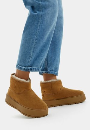 Bottes en daim marron à hauteur de cheville avec une doublure en polaire douce, au design arrondi et à semelles en caoutchouc épaisses, associées à un jean léger bleu clair retourné.