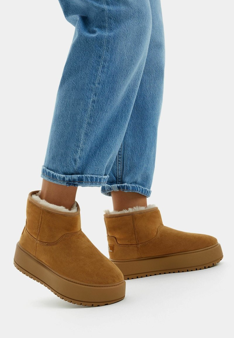 Brown suede ankle boots with a soft fleece lining, rounded design, and thick rubber soles, paired with cuffed light blue jeans.
