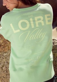 Mündroheline kapuutsiga sweatshirt, millel on julge valge kiri "THE LOIRE Valley" ning lisainfot piirkondade ja viinamarjade kohta.
