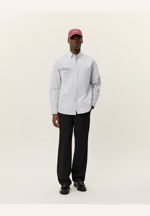 Gestreepte button-up shirt in lichtblauw en wit, gecombineerd met zwarte broek en een bordeauxrode pet met de tekst "Lower West Side". Glad stof.