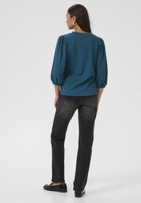 Blouse en teal texturé avec des manches bouffantes, associée à un jean noir droit et des mocassins noirs brillants. Design minimaliste, vue arrière.