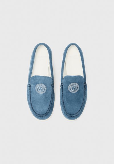Zilā suede kurpes ar noapaļotu purngalu, kas ir aprīkotas ar dekoratīvām šuvēm un apaļu logotipa emblēmu augšējā daļā.