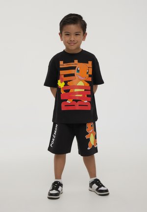 T-shirt nera a maniche corte con grafica di Charmander, dettagli arancioni sul petto e sulla schiena. Pantaloni corti neri con design di Charmander. Sneakers bianco e nere.