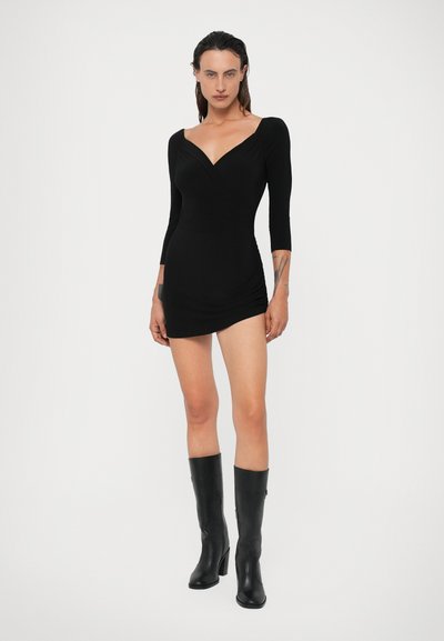 Černé dlouhé šaty s výstřihem do V a staženým pasem, s přiléhavým siluetou. Nosené s černými kozačkami na podpatku po kolena.