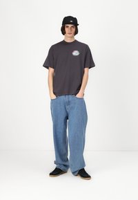 Mørk grå t-skjorte med et rundt logo på venstre side, parret med løse blå jeans og svarte joggesko. Capsen er iført bakpå, flatt design.