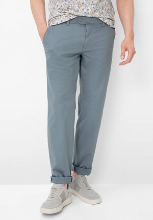 Uomo che indossa pantaloni grigi risvoltati, sneakers grigio chiaro e una camicia fantasia con le mani parzialmente nelle tasche, in piedi contro uno sfondo bianco.