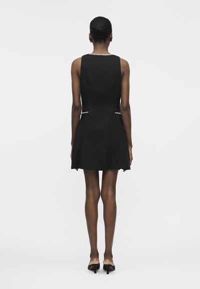 Γυναίκα με σκούρο δέρμα που φοράει μαύρο αμάνικο φόρεμα με λευκή διακόσμηση, όρθια και στραμμένη προς τα πίσω, φοράει μαύρα παπούτσια με τακούνι.