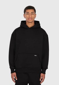 Jeune homme portant un sweat à capuche noir uni avec une poche frontale, se tenant devant un fond gris clair, faisant face à l'objectif avec une expression neutre.