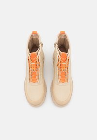 Beżowe sneakersy za kostkę z pomarańczowym akcentem, chropowata tkanina jako górna część, wyraźny detal sznurowania. Gruba gumowa podeszwa, funkcjonalny zamek błyskawiczny.