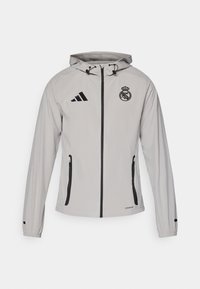 Lys grå hættejakke med sort lynlås, sidelommer, Adidas-logo på venstre bryst og Real Madrid-crest på højre bryst.