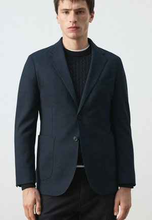 Young man wearing a dark navy blazer over a black cable-knit sweater and black pants, standing against a plain light background.