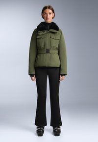 Olivno zelena puffer jakna z krznom obloženim ovratnikom, dvema sprednjima žepoma, pasom na voljo, v kombinaciji z črnimi širokimi hlačami in masivnimi črnimi čevlji.