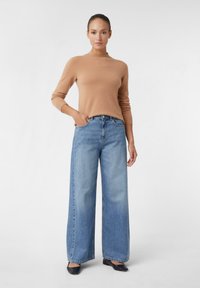 Frau steht mit einer Hand in der Tasche, trägt ein beiges Langarmoberteil, helle, weite Jeans und schwarze spitze Ballerinas.
