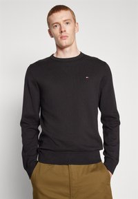 Black long-sleeve sweater with a ribbed texture and crew neckline, featuring a small logo on the chest. Paired with light brown trousers.