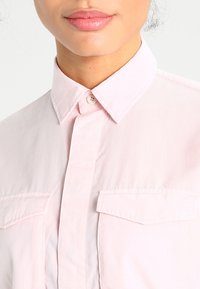 Camicia rosa chiaro realizzata in un tessuto dalla consistenza liscia. Presenta un colletto a punta, chiusura con bottoni e due tasche sul petto.