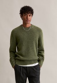 Πράσινη ζακέτα από υλικό υφασμάτινης πλέξης, με στρογγυλή λαιμόκοψη, ριμπ τελείωμα και κίτρινες πινελιές παντού. Φορεμένη πάνω από ένα λευκό πουκάμισο.