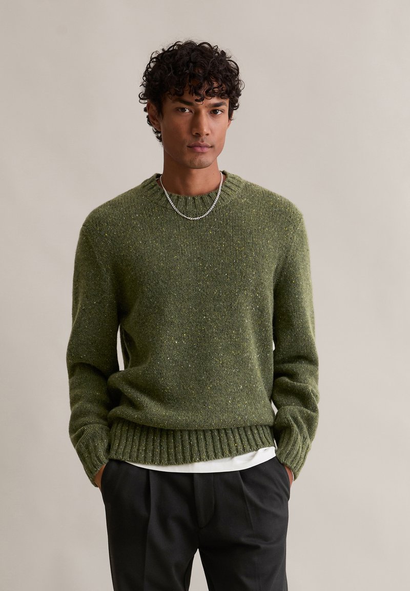 Πράσινη ζακέτα από υλικό υφασμάτινης πλέξης, με στρογγυλή λαιμόκοψη, ριμπ τελείωμα και κίτρινες πινελιές παντού. Φορεμένη πάνω από ένα λευκό πουκάμισο.