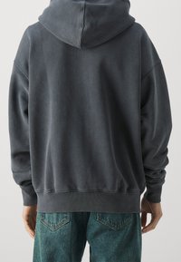 Persona che indossa un felpa grigia scura e jeans di denim blu, vista da dietro con le mani rilassate ai fianchi.