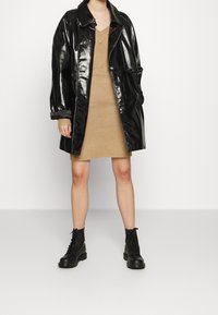 Trench-coat noir brillant porté sur une robe-pull camel, assorti à des bottes de combat noires montantes jusqu'aux genoux. Doté d'un col et de poignets retroussés.