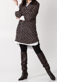 Czarna tunika w kwiatowy wzór, nałożona na białą koszulę, w połączeniu z czarnymi rajstopami i brązowymi skórzanymi butami za kolano.