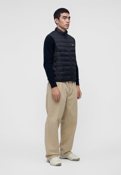 Homme debout face à droite portant un gilet matelassé noir, une chemise noire à manches longues, un pantalon large beige et des baskets blanches sur fond blanc.