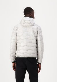 Giacca a vento argentata con motivo trapuntato, cappuccio e polsi a coste. Il tessuto ha una finitura lucida, offrendo isolamento e calore.