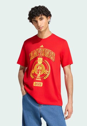 Junger Mann mit lockigem Haar, der ein rotes "Campeones 2026" T-Shirt mit einer goldenen Pokal-Grafik und blauen Jeans trägt, steht mit einer Hand in der Tasche.