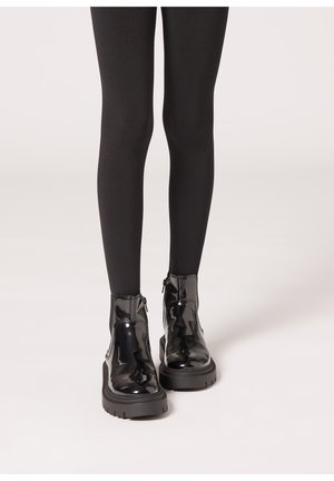 Sorte patent læder ankelstøvler med en tekstureret overflade, kraftig gummisål og lynlås i siden, parret med sorte leggings.
