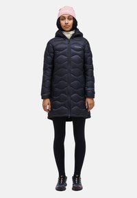 Black puffer coat with a wavy quilted pattern, zip closure, hood, and two front pockets. Worn over black leggings and hiking shoes.