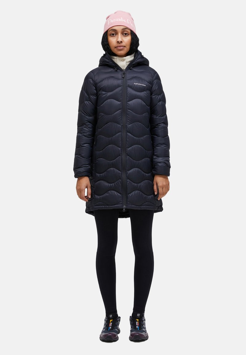 Black puffer coat with a wavy quilted pattern, zip closure, hood, and two front pockets. Worn over black leggings and hiking shoes.