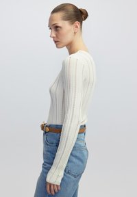 Weißes langärmliges Strickoberteil mit strukturierten vertikalen Streifen, kombiniert mit hoch taillierten blauen Jeans und einem braunen Ledergürtel.