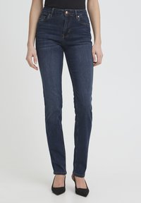 Femme portant un jean slim bleu foncé et des talons noirs à bout pointu, debout devant un fond clair uni.