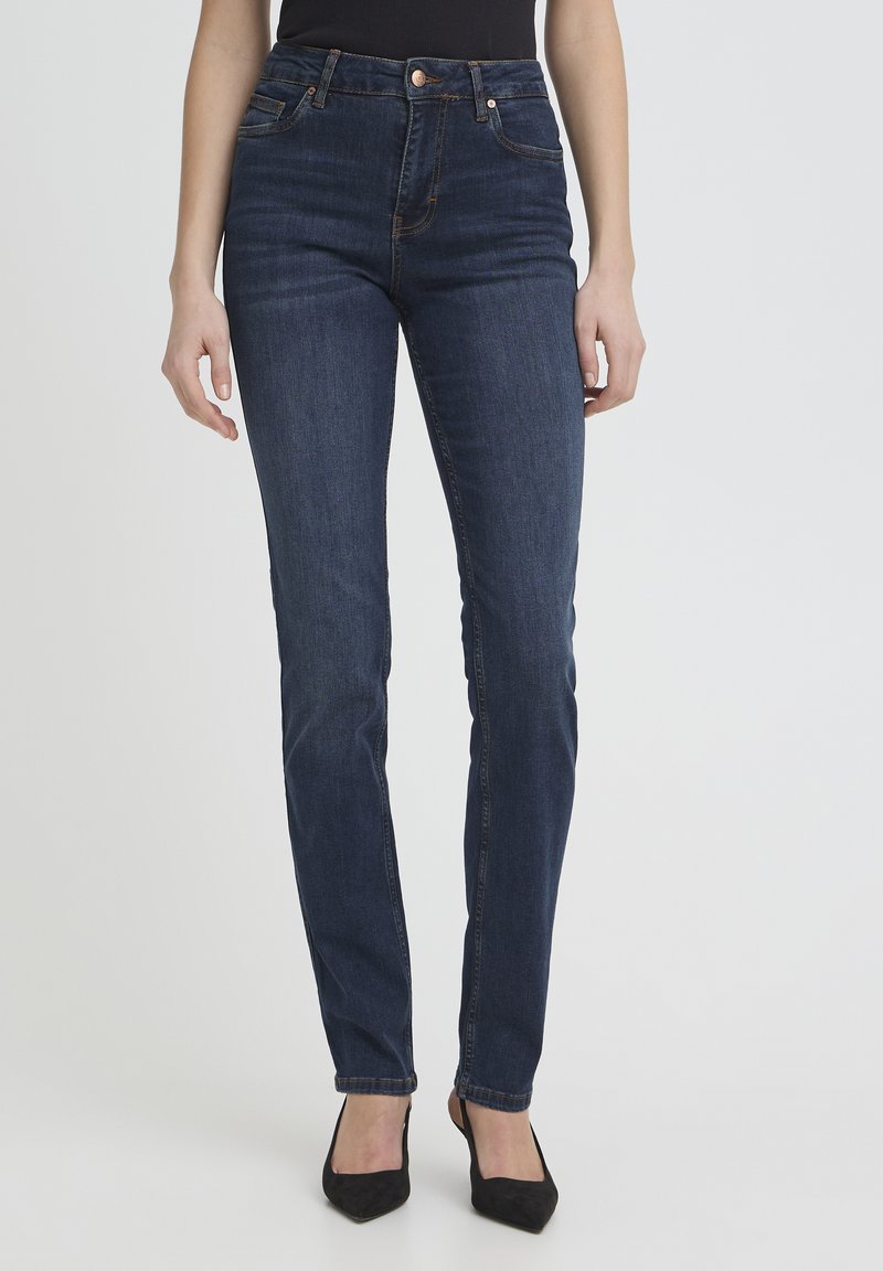 Femme portant un jean slim bleu foncé et des talons noirs à bout pointu, debout devant un fond clair uni.