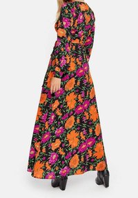 Robe maxi avec une base noire ornée de motifs floraux vibrants en orange et rose. Manches longues et silhouette fluide. Bottes à talons noires.