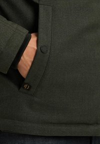 Dark green jacket fabric with a textured finish, featuring a side pocket, button detail, and a small label at the hem.