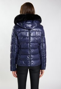 Giacca puffer blu navy con cappuccio foderato in pelliccia, design trapuntato, finitura lucida e dettagli nelle cuciture laterali. Caratterizzata da maniche lunghe e una silhouette aderente.