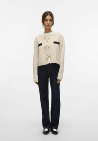 Crème gebreide cardigan met zwarte zakaccenten en strikdetails, gecombineerd met donkerblauwe flared jeans en zwarte schoenen met witte rand.