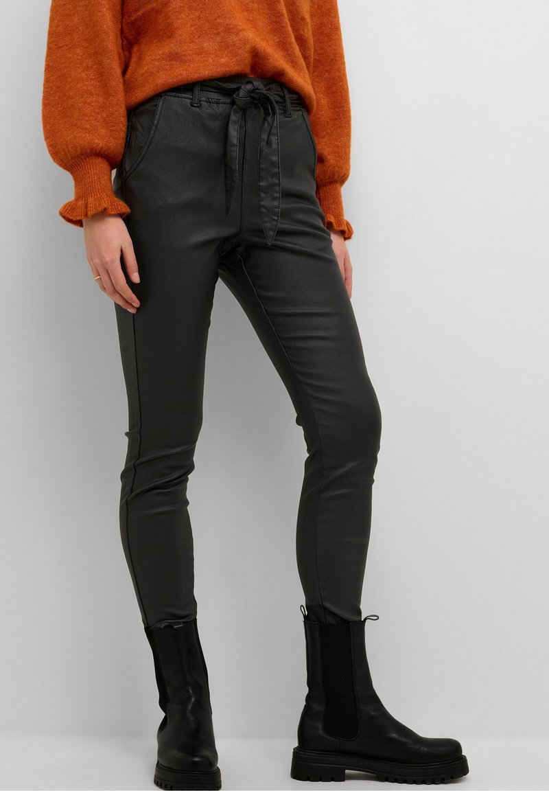 Pantalon en similicuir noir à taille haute avec un détail noué, associé à un pull orange et des bottines noires, présentant un tricot texturé.