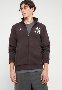Mlad moški nosi temno rjavo jakno na zadrgo z logotipom New York Yankees, sivo majico in sive črtaste hlače, gleda levo.