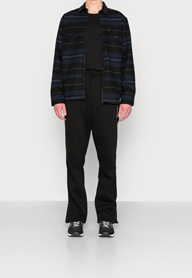 Czarna i niebieska kurtka w paski z dwoma kieszeniami na piersi, zestawiona z czarnymi spodniami i sportowymi butami. Gładki materiał i luźny krój na całej długości.
