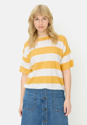 Blondynka w żółto-białej, paskowanej koszuli z krótkim rękawem i zapinanej na guziki niebieskiej spódnicy dżinsowej, stojąca na tle jednolitego tła.