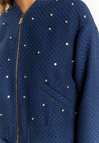 Marineblauwe gebombardeerde jas met textuur, voorzien van een ritssluiting aan de voorkant, bezaaid met gouden studs en een voorzak. Dit biedt een gestructureerd, modern silhouet.