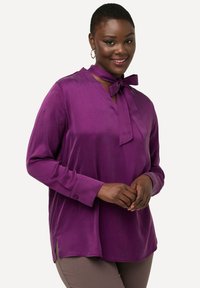 Paarse satijnen blouse met lange mouwen, een V-halslijn en een strikdetail bij de kraag. Heeft zijsplitten aan de zoom.