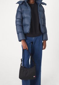 Une personne portant une doudoune marine et un jean bleu tient un sac à bandoulière noir avec un petit logo blanc et rouge.