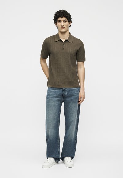 Νέος άνδρας με σγουρά μαλλιά στέκεται φορώντας καφέ κοντομάνικο πλεκτό πόλο, φαρδιά μπλε τζιν και λευκά αθλητικά σε απλό φόντο.