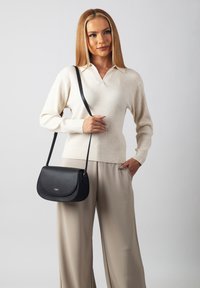 Black leather shoulder bag with a curved flap, subtle stitching, and a detachable strap, worn over a cream sweater and beige trousers.