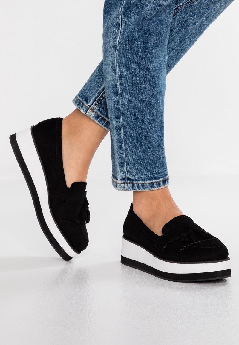 Pieds portant des mocassins plateforme en daim noir avec des glands et des semelles blanches, assortis à un jean bleu retroussé.